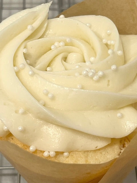 A close up of the beautifully swirled vanilla frosting dotted with tiny pearl white nonpareils. the vanilla cupcake peaks through. The frosting glistens.