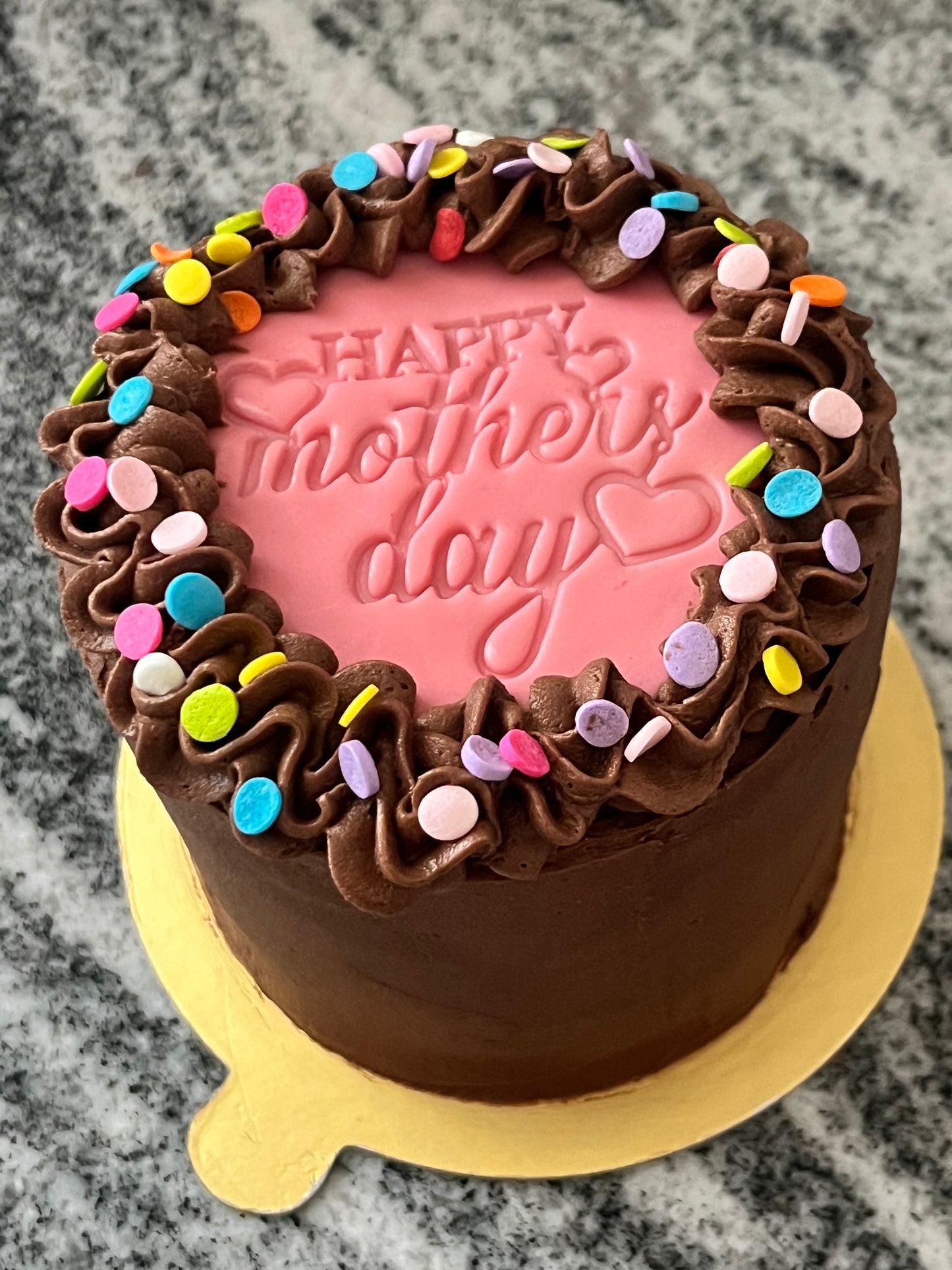 The top side view of a chocolate mini cake with chocolate frosting, a pink ‘Happy Mother’s Day’ marshmallow fondant topper and multi-colored confetti.