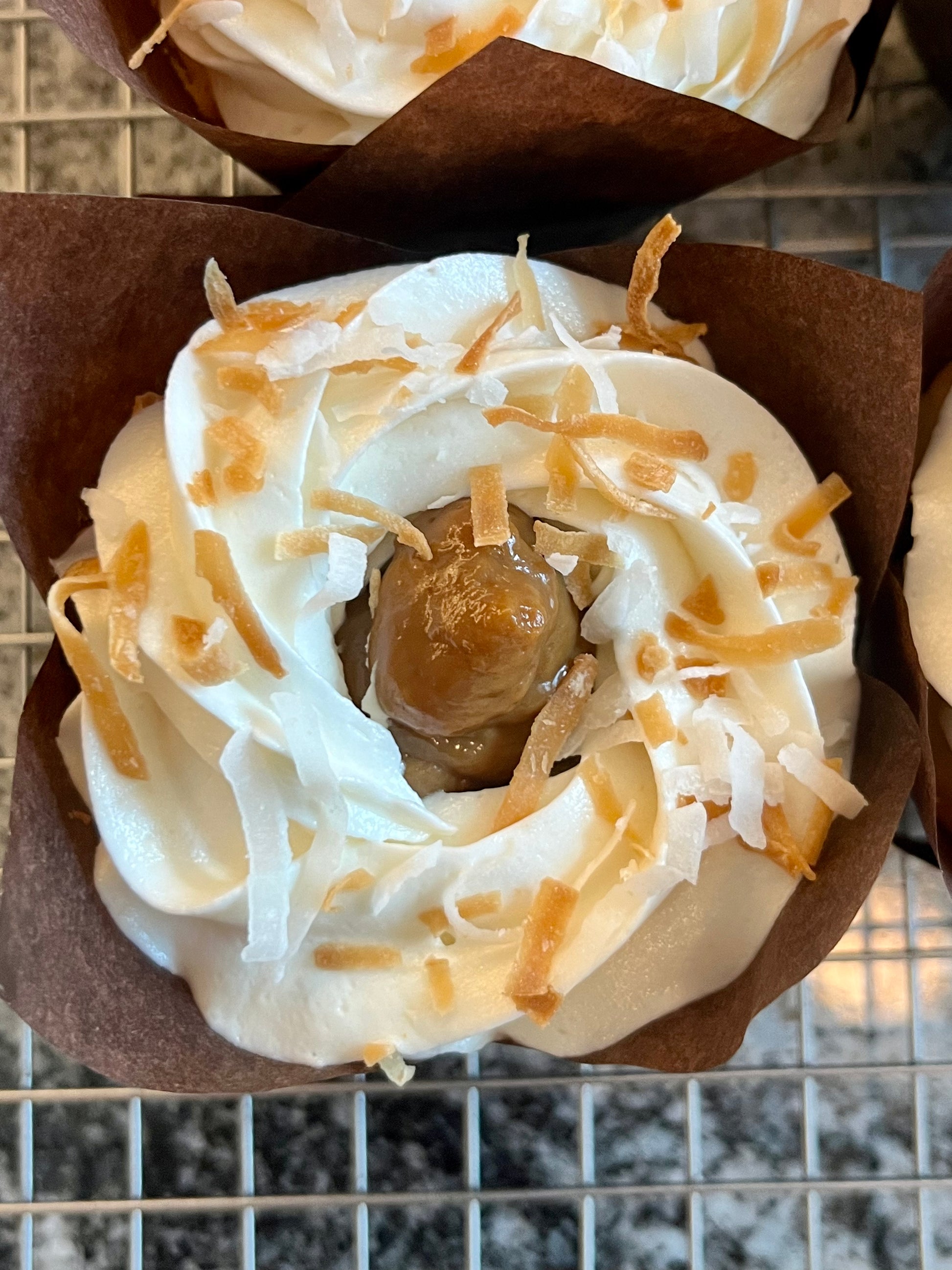 A close up of the top of a Coconut Dulce de Leche cupcake crowned with a decadent swirly ribbon of coconut buttercream topped with toasted coconut. The Dulce de Leche peaks through the buttercream swirl.