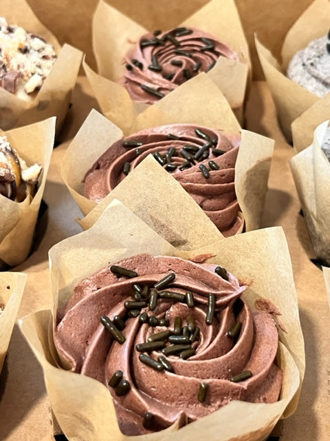 Three Chocoholic cupcakes sit in a bakery box.  The chocolate cupcakes are topped with a ribboned swirl of chocolate fudge frosting and crowned with chocolate sprinkles.