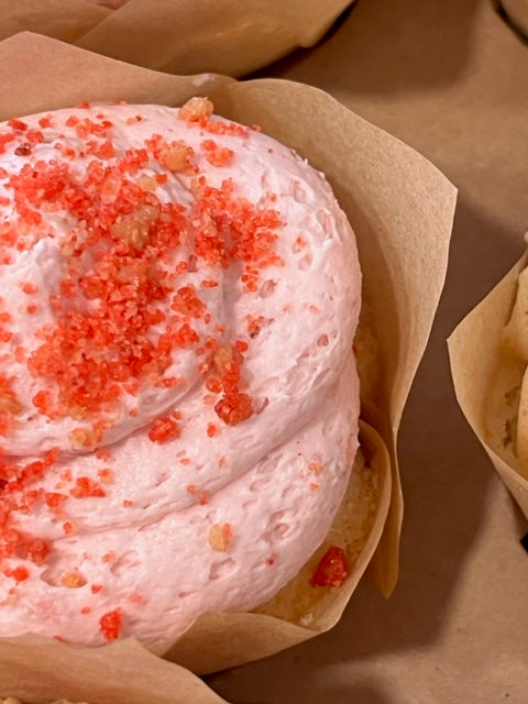 Close up of a Strawberry Milkshake cupcake.  The vanilla cupcake peeks out from under a strawberry buttercream frosting swirl topped with speckles of strawberry crumbs.