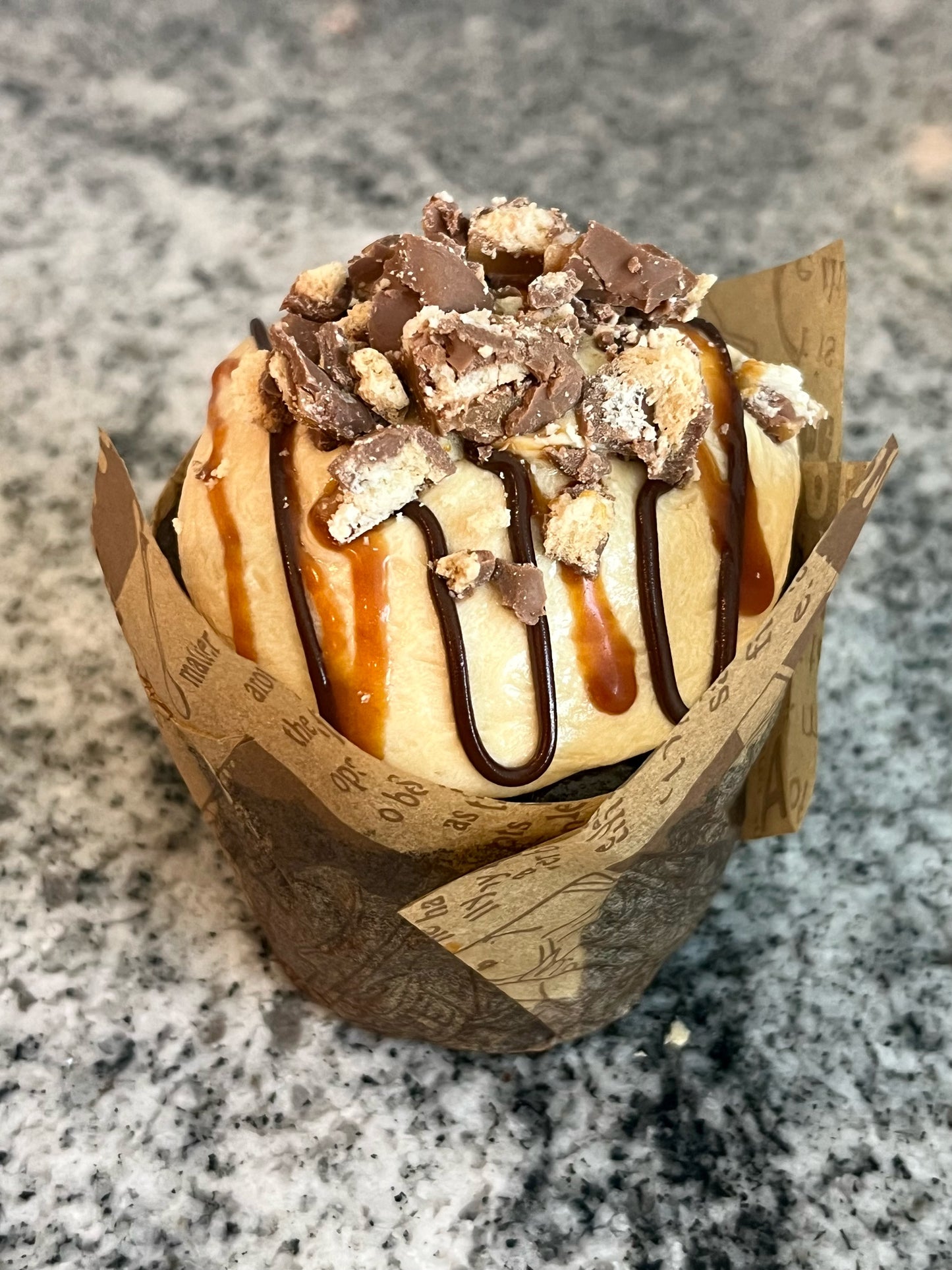 a close up of a single Chocolate Salted Cupcake. The frosting glistens, drizzled with homemade chocolate fudge and salted caramel , crowned with Twix candy chunks.