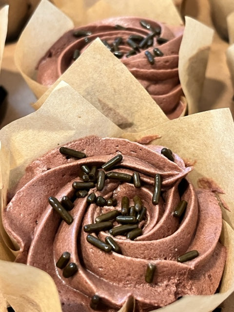 A close up of two Chocoholic cupcakes in a bakery box.  The chocolate cupcakes are topped with a ribboned swirl of chocolate fudge frosting and crowned with chocolate sprinkles.