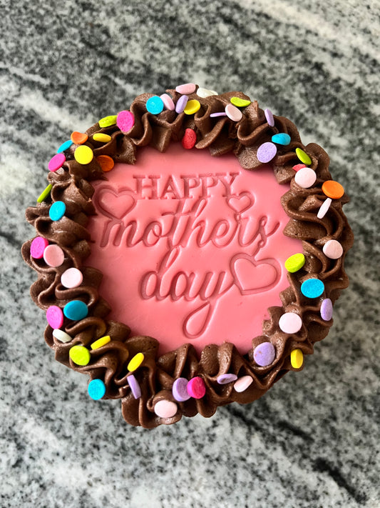 The top view of a chocolate mini cake with chocolate frosting, a pink ‘Happy Mother’s Day’ marshmallow fondant topper and multi-colored confetti.