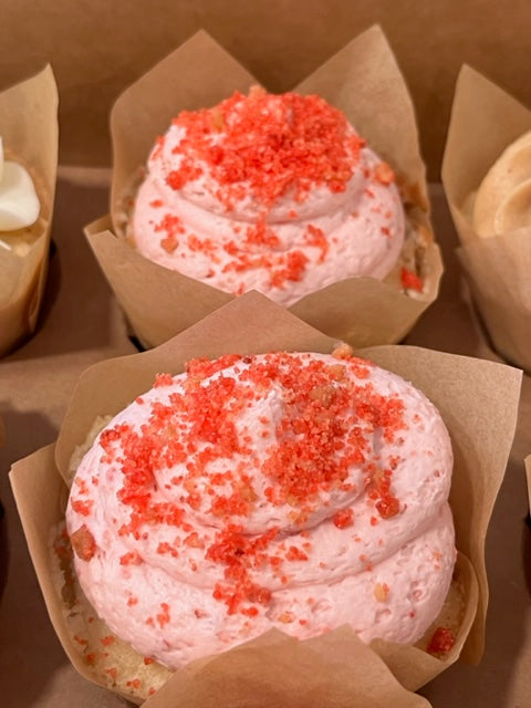 Two Strawberry Milkshake cupcakes rest in a bakers box.  On the middle cupcake, the vanilla cake peeks out from under a strawberry buttercream frosting swirl topped with speckles of strawberry crumbs.