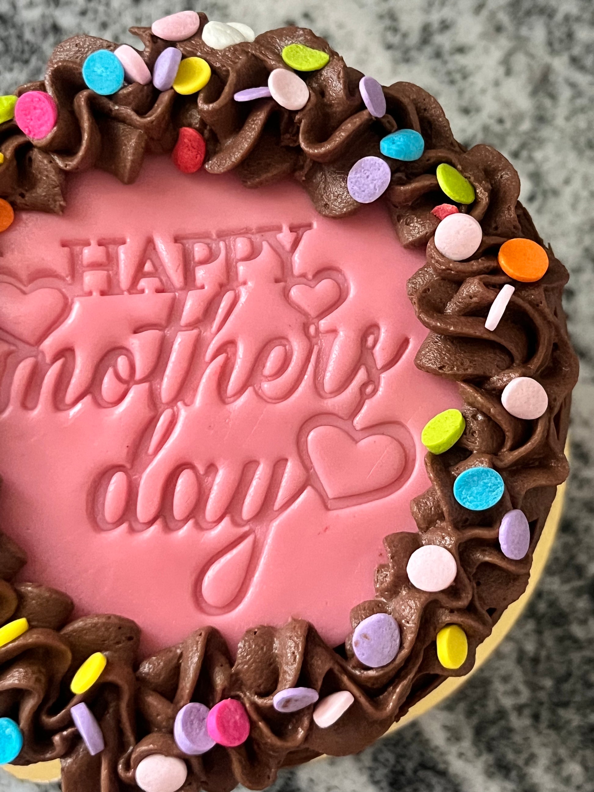 A close up of the top of a chocolate mini cake with chocolate frosting, a pink ‘Happy Mother’s Day’ marshmallow fondant topper and multi-colored confetti.