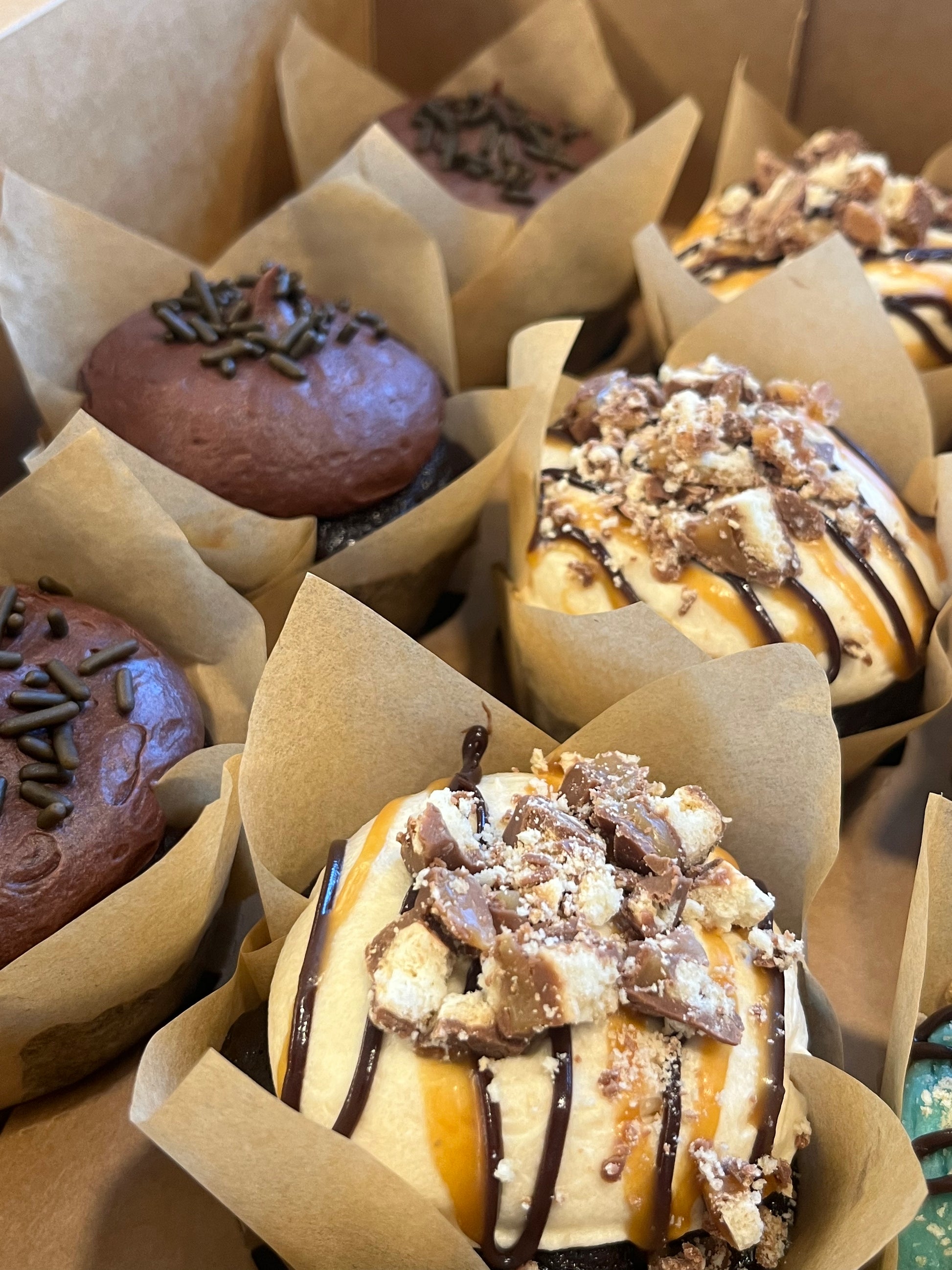 Three Chocolate Salted Caramel cupcakes sit in a bakers box next to three Chocoholic cupcakes.  The frosting glistens, drizzled with homemade chocolate fudge and salted caramel , crowned with Twix candy chunks.