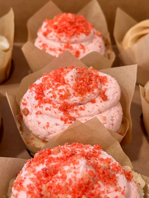 Three Strawberry Milkshake cupcakes rest in a bakers box.  On the middle cupcake, the vanilla cake peeks out from under a strawberry buttercream frosting swirl topped with speckles of strawberry crumbs.