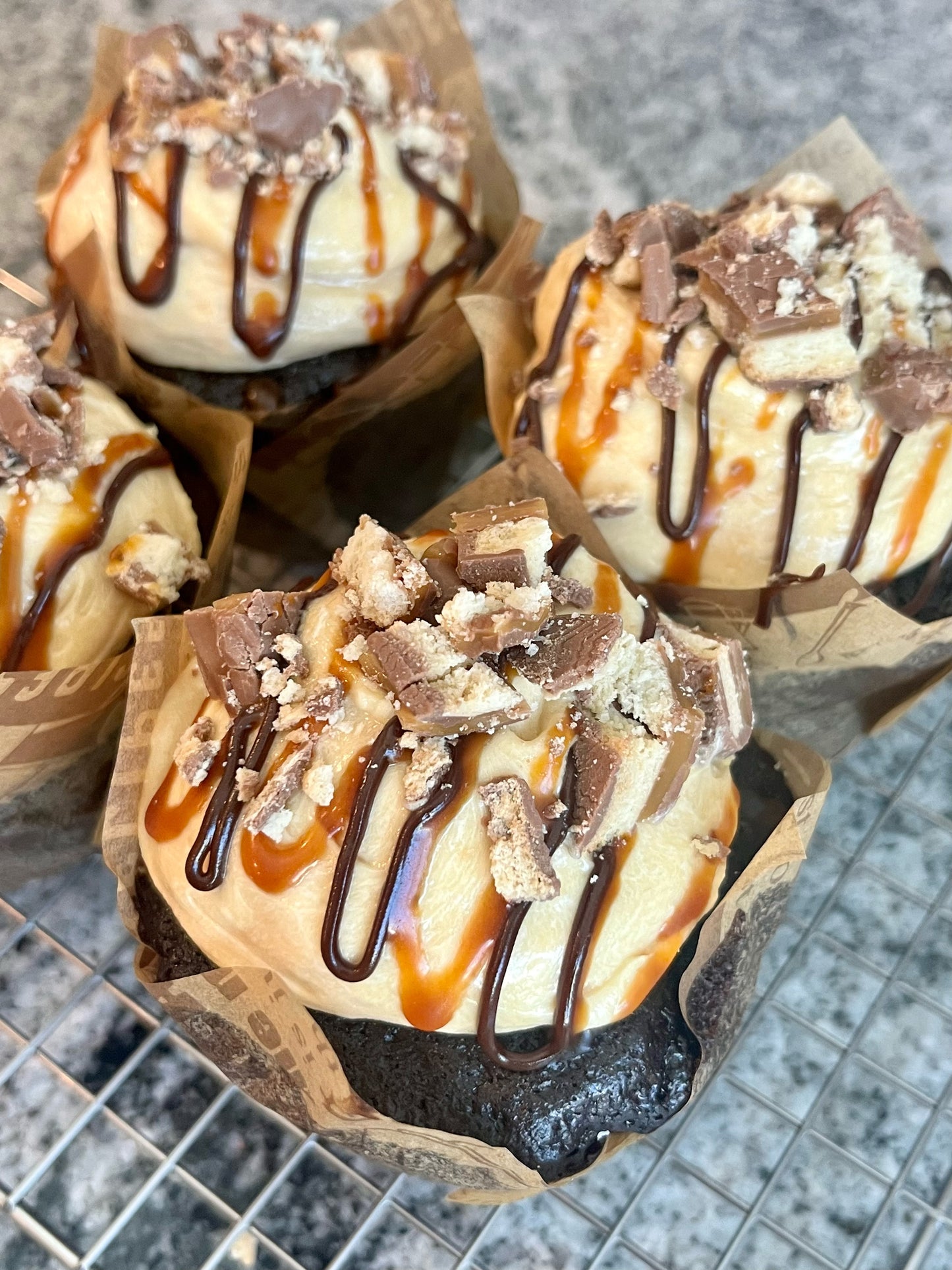 Four Chocolate Salted Caramel cupcakes sit on a bakers rack.  The frosting glistens, drizzled with homemade chocolate fudge and salted caramel , crowned with Twix candy chunks.