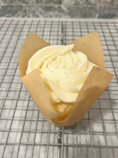 A Very Vanilla cupcake sits alone on a bakery rack.  The vanilla cupcake peaks through the small gap between the wrapper and the frosting.