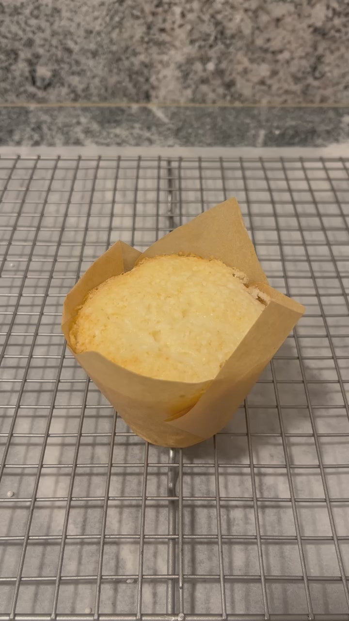 A vanilla cupcake in a light brown tulip wrapper sits atop a baking rack.  A gorgeous, rippled swirl of frosting is then piped onto a vanilla cupcake and tiny pearl white nonpareils are sprinkled on top.